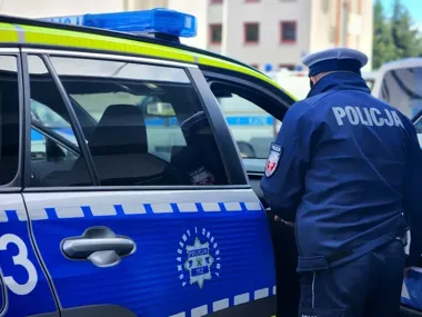 Policjant w mundurze stoi przy otwartych drzwiach oznakowanego radiowozu policyjnego na tle budynków.
