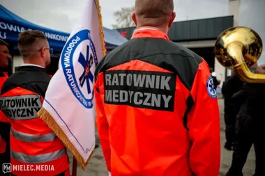 Ratownicy medyczni w pomarańczowo-czarnych kurtkach stoją podczas uroczystości, jeden z nich trzyma sztandar z napisem „Podgórze Ratunkowego w Mielcu”.
